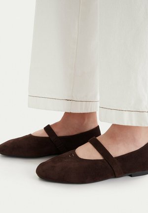 Dunkelbraune Mary Jane Ballerinas aus Wildleder, getragen zu weißen, gekürzten Hosen, mit einem einzelnen Riemen über dem Fußspann und flachen schwarzen Sohlen.