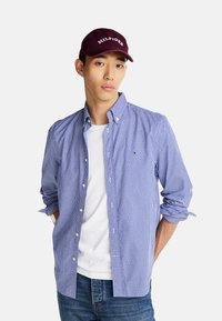 Camisa de cuadros azul y blanca de manga larga con cuello abotonado, llevada sobre una camiseta blanca, combinada con unos vaqueros de mezclilla y una gorra burdeos.