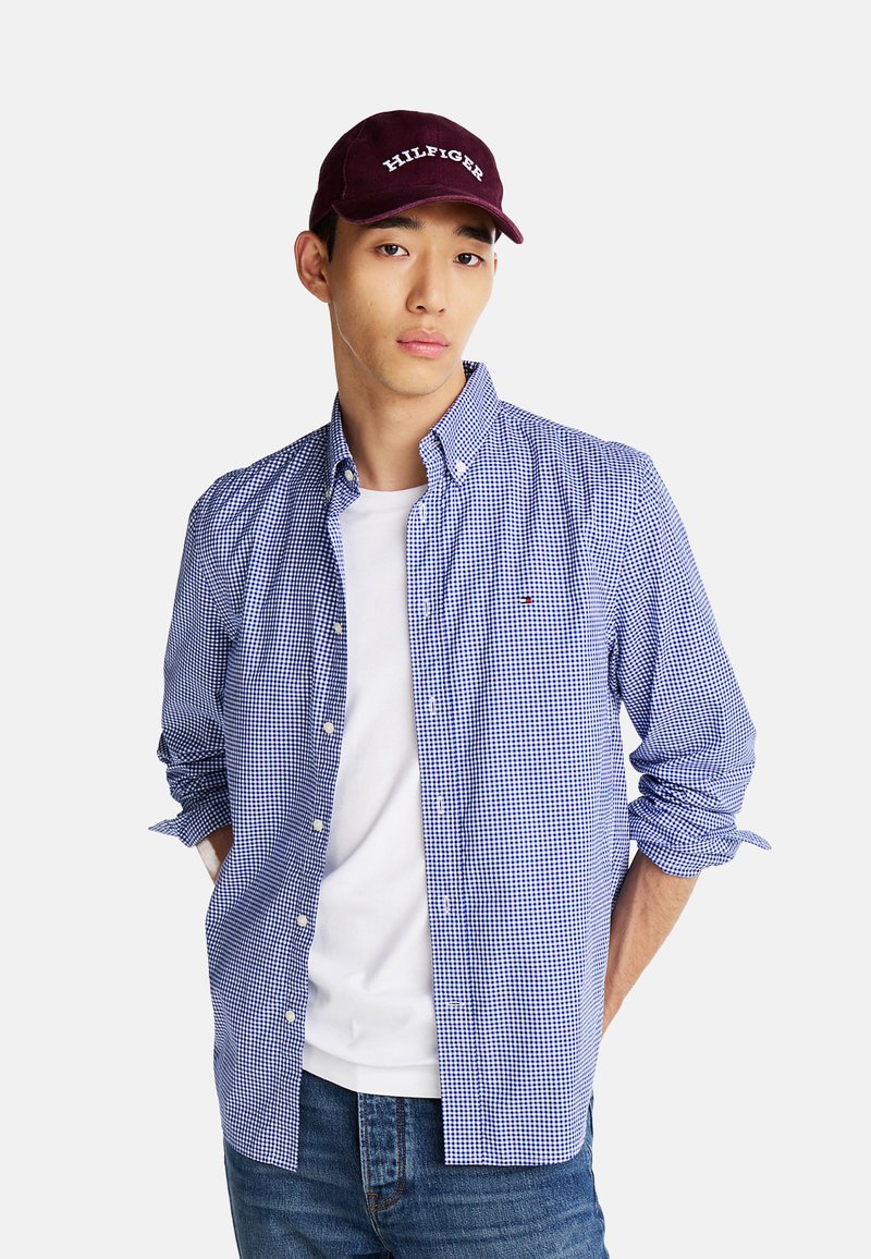 Camisa de cuadros azul y blanca de manga larga con cuello abotonado, llevada sobre una camiseta blanca, combinada con unos vaqueros de mezclilla y una gorra burdeos.