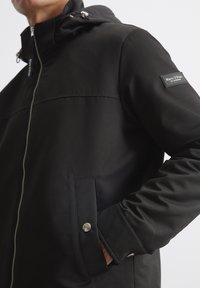 Czarna bluza zapinana na zamek wykonana z gładkiej tkaniny, z kapturem, przednimi kieszeniami na zatrzaski oraz naszywką z logo na rękawie.