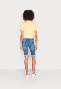 Lyhyet siniset farkkushortsit, joissa on käärityt lahkeet ja takataskut, yhdistettynä keltaiseen lyhytpaitaan ja valkoisiin lenkkareihin, joissa on punaisia yksityiskohtia.