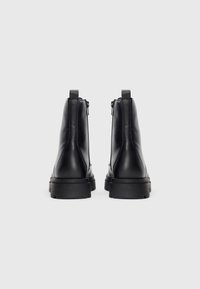 Une paire de bottes noires en cuir à cheville avec de épaisses semelles en caoutchouc, présentée de derrière sur un fond blanc uni.
