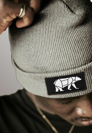 Person adjusting light gray knit beanie with a black patch featuring a geometric white bear design.