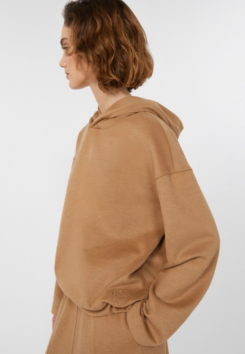 Frau mit kurzem, welligem Haar, die einen lockeren, beigen Kapuzenpullover und passende Hose trägt, im Profil vor einem schlichten weißen Hintergrund abgebildet.