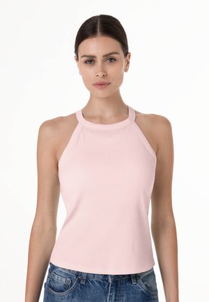 Giovane donna con capelli scuri, indossa un top rosa chiaro senza maniche a coste e jeans blu, in piedi davanti a uno sfondo bianco uniforme.