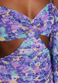 Blomstret kjole med et mønster i lilla, gult og grønt. Materialet er tekstureret med rynker og udskæringsdetaljer nær taljen.