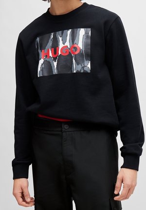 Persoon draagt een zwarte trui met een grijs-wit abstract patroon en rode "HUGO" tekst, gecombineerd met een zwarte broek.