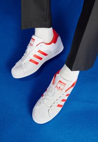 Zapatillas deportivas blancas con acentos rojos y tres rayas, con una puntera texturizada y un logo prominente en la lengüeta, acompañadas de calcetines blancos.