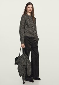 Cardigan lavorato a maglia nero e crema con bottoni, abbinato a pantaloni scuri a gamba larga. Il modello tiene in mano un cardigan abbinato e una borsa nera.