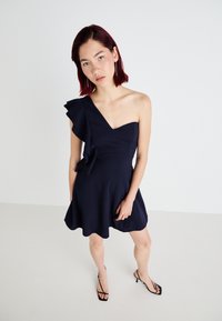 Vestido azul-marinho de um ombro só com detalhes em babados, corpete justo e saia rodada, confeccionado em um tecido suave. Sandálias pretas de tiras. 