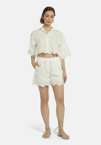 Witte eyelet crop top met gegolfde rand, gecombineerd met bijpassende witte shorts met een elastische tailleband en zakken, beide met een patroon textuur.