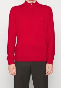 Pull-over en tricot rouge avec un col montant et une demi-fermeture éclair, doté de manches longues et d'un logo discret sur la poitrine. Porté avec un pantalon noir.