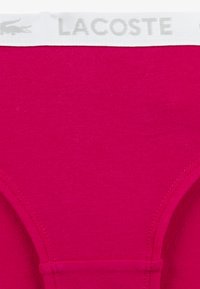 Bright pink fabric with a ribbed texture. White waistband features the brand name "LACOSTE" in a subtle gray print.