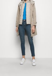 Kvinna klädd i en beige lagerjacka, ljusblå topp, åtsittande blå jeans och vita sneakers som står framför en vit bakgrund.