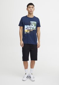 Camiseta de algodón azul marino con un estampado gráfico de palmeras y el texto "MEZCLA DE VERANO INFINITO". Combinada con pantalones cortos negros y zapatillas blancas.