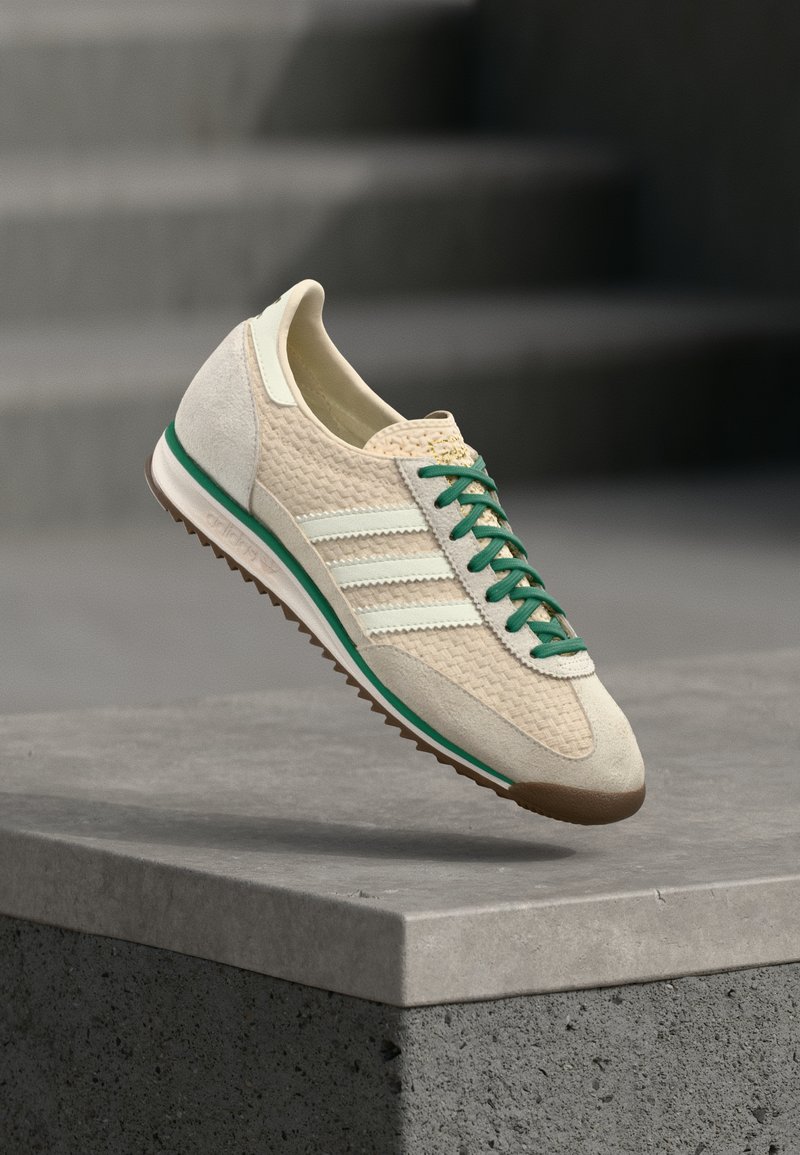 Μπεζ αθλητικό παπούτσι με υφασμάτινο πλέγμα και σουέντ λεπτομέρειες, πράσινα κορδόνια, λευκές ρίγες και καφέ λαστιχένια σόλα. Κλασικός αθλητικός σχεδιασμός.