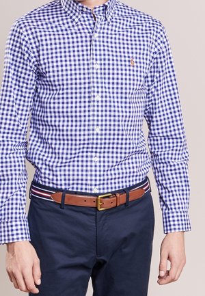 Mężczyzna w niebiesko-białej koszuli w kratę, włożonej do granatowych spodni z brązowym skórzanym paskiem w czerwono-biało-niebieskie paski.