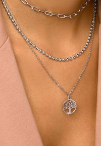 Tre collane d'argento sovrapposte: una con un pendente a forma di albero circolare, un'altra con perle tonde e una terza con maglie allungate.