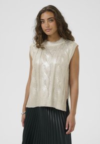 Neuletta ilman hihoja kimaltelevassa kermassa, jossa on palmikkokuvio, korkealla kauluksella ja sivurei'illä, yhdistettynä pleatattuun mustaan hameeseen.