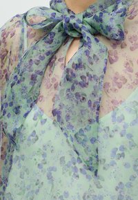 Blouse transparente à motif floral violet sous une écharpe verte à fleurs nouée en nœud à l'encolure.