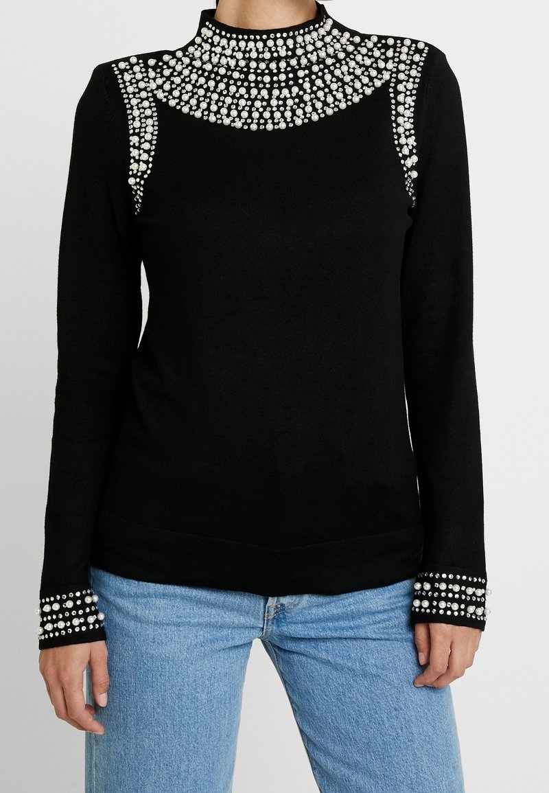 Schwarzer Pullover mit hohem Kragen, verziert mit Strasssteinen an den Schultern und Ärmeln. Weiches Material, taillierte Passform, lange Ärmel und strukturierte Akzente.