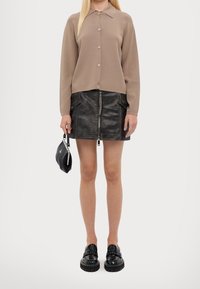 Beżowy sweter na guziki z długimi rękawami, zestawiony z czarną mini spódnicą zapinaną na zamek z czarnej skóry. Modelka trzyma czarną torebkę i nosi czarne buty.