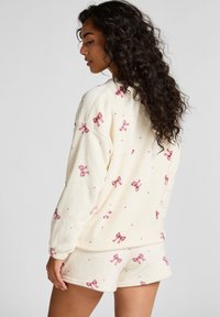 Crème fleece loungewear set met roze strikontwerpen, langemouwen top met elastische manchetten, en bijpassende korte broek met vergelijkbare patronen.