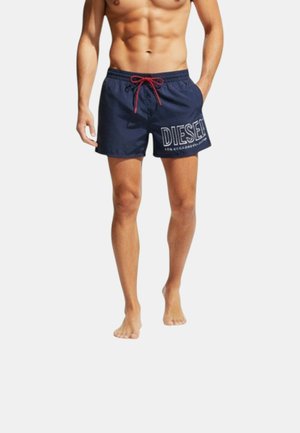 Hombre sin camiseta con bañador Diesel azul marino con cordón rojo, de pie descalzo frente a un fondo blanco liso.