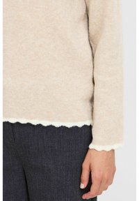Beige ulltröja med en vågig vit nederkant och manschetter, mjukt material, i kombination med mörkgrå byxor med en strukturerad yta.