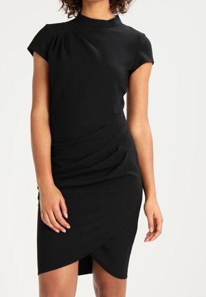 Mujer con un vestido negro ajustado de manga corta, cuello alto y falda asimétrica, de pie sobre un fondo claro y liso.