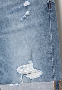 Tissu en denim bleu clair avec des zones usées et des trous effilochés, laissant apparaître des fils blancs en dessous, et un ourlet plié avec une couture marron.