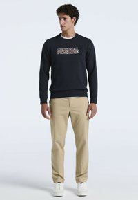Homme debout portant un sweat-shirt marine avec le texte "Original Penguin", un pantalon beige et des baskets blanches, regardant légèrement sur sa gauche.