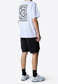 Biała bawełniana koszulka z nadrukiem na plecach, w komplecie z czarnymi szortami cargo i jasnoszarymi sneakersami.