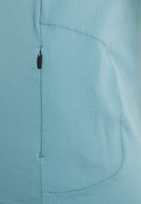 Jasnoniebieska tkanina o gładkiej fakturze. Posiada kieszeń na suwak z boku oraz subtelne detale szwów. Całość zaprojektowana bezszwowo.