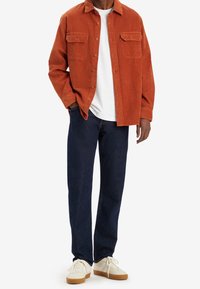 Orange kordflorskjorta med två bröstfickor, bärs över en vit t-shirt. Matchad med mörkblå byxor och krämfärgade sneakers med gummiklädda sulor.