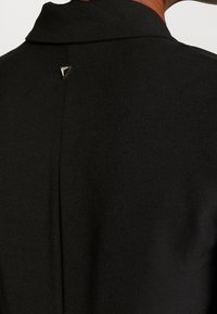 Abrigo negro a medida, con una textura suave, un cuello bajo y un acento metálico triangular en la parte posterior del cuello. Líneas limpias y un diseño discreto.