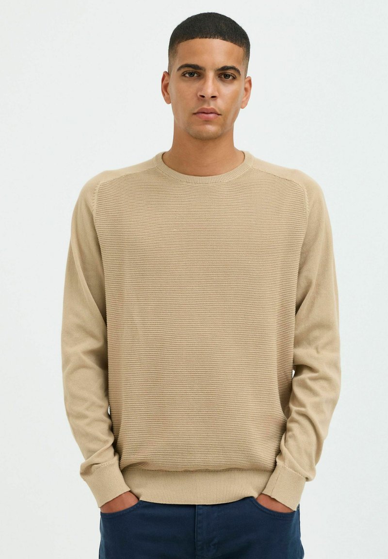 Pull en tricot beige avec une texture côtelée, un col rond et des manches longues. Présente une coupe décontractée et des rayures horizontales.