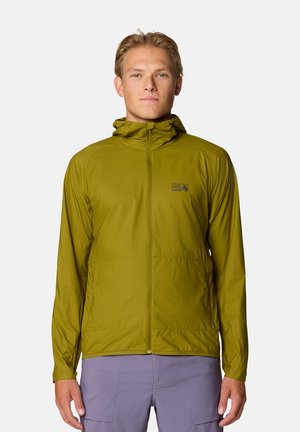 Junger Mann trägt eine olivgrüne Kapuzenjacke mit Reißverschluss und hellviolette Hose, steht vor einem schlichten hellen Hintergrund.