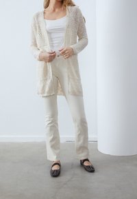 Žena v krémovém krajkovém kardiganu, bílém tílku, krémových kalhotách a černých balerínách, stojící na betonové podlaze vedle bílé kolony.