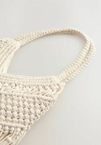 Gehaakte macramé tas in gebroken wit, met gevlochten bandjes, een gestructureerd oppervlak en decoratieve kralen langs de rand.