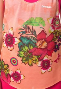 Brzoskwiniowy top bez rękawów z wielokolorowymi wzorami kwiatów i liści, w tym czerwonych, zielonych i żółtych kwiatów, noszony na jasnoróżowej odzieży.