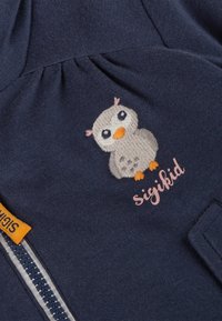 Dunkelblauem Stoff mit einem gestickten Cartoon-Eulen-Motiv mit orangefarbenem Schnabel und Füßen, neben dem Text "sigikid" und einem teilweise sichtbaren Reißverschluss.