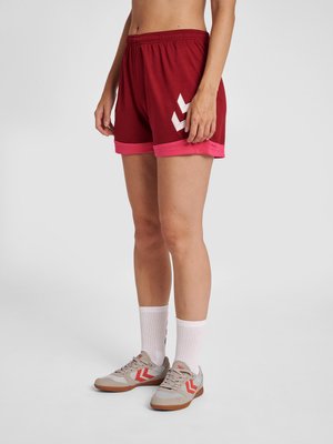 Person trägt rote Sportshorts mit pinkem Besatz, weiße Sportsocken und beige Sneakers mit roten Chevron-Logos und steht vor weißem Hintergrund.