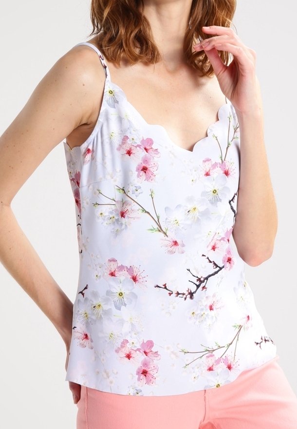 Woman wearing a white sleeveless top with scalloped neckline and pink and white floral print, paired with light pink pants.