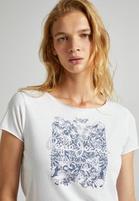 Camiseta blanca de manga corta con un patrón floral azul y el texto "Pepe Jeans LONDON" impreso en el centro. Material de algodón suave.