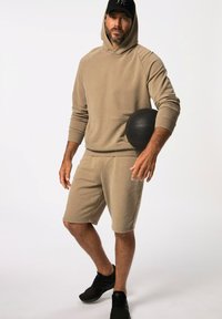 Felpa beige con tasca frontale e pantaloni corti coordinati, caratterizzati da una texture liscia. Palla da esercizio nera tenuta sotto il braccio. Sneakers nere.