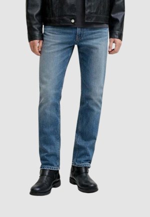 Uomo che indossa jeans aderenti azzurri con aree sbiadite, giacca di pelle nera e stivaletti neri, in piedi contro uno sfondo semplice.