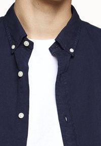 Homme portant une chemise bleu marine déboutonnée sur un t-shirt blanc à col rond, avec les boutons du col fermés.