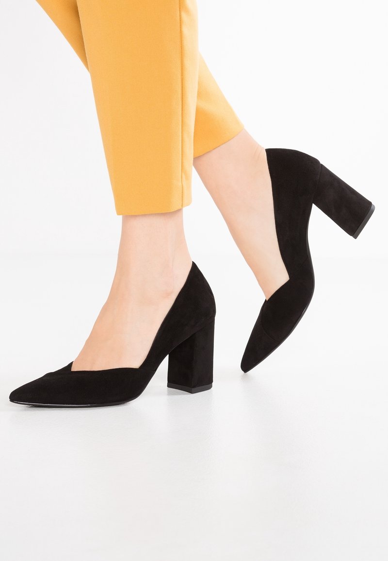 Femme portant un pantalon jaune moutarde et des escarpins noirs en daim à talon bloc, un pied légèrement levé sur un fond blanc.