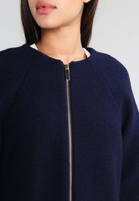 Giacca in misto lana blu navy con colletto rotondo, zip frontale dorata e texture a maglia con maniche raglan. Nessun accento o motivo aggiuntivo.